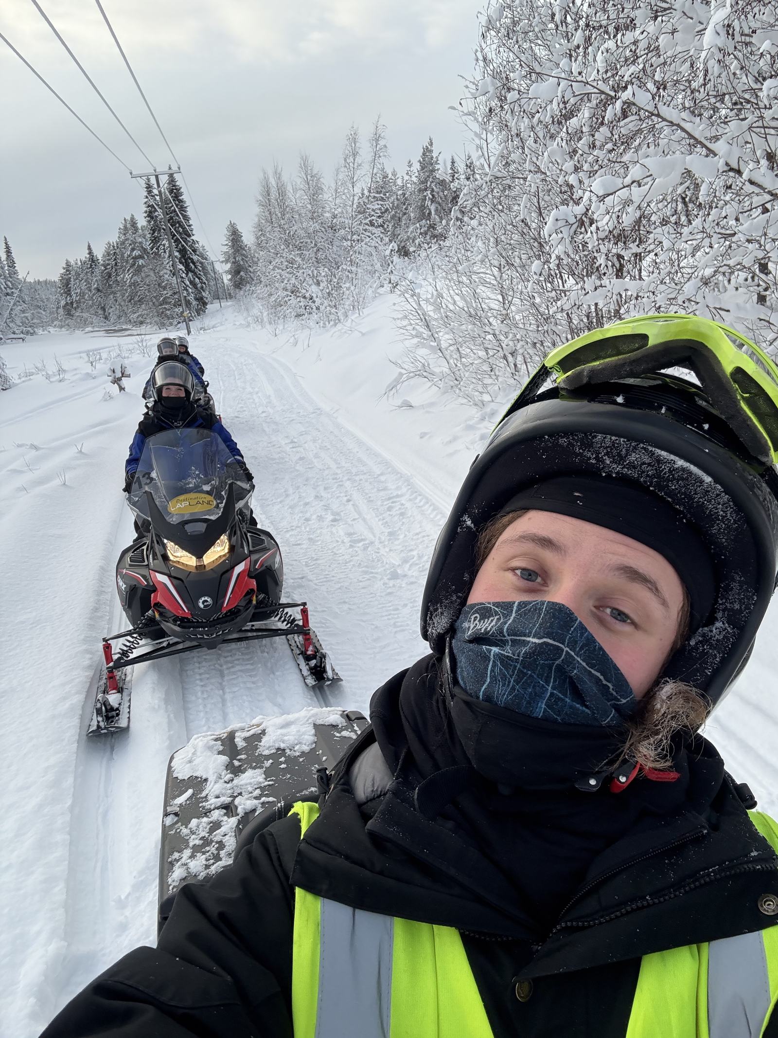 Guide-Selfie auf dem Schneemobil, dahinter die Gruppe im verschneiten Wald
